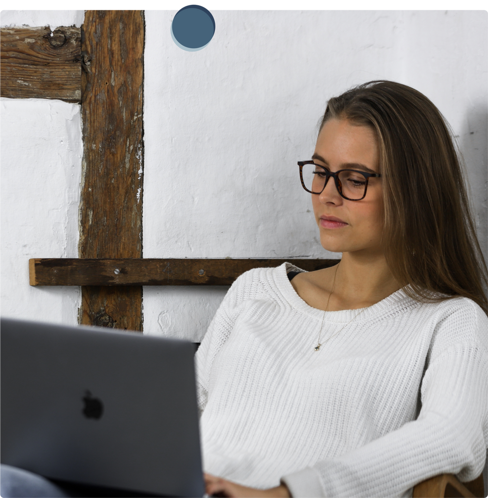 Eine jüngere Frau mit weißem Pullover trägt eine Blaufilter Brille, während Sie auf den Laptop schaut